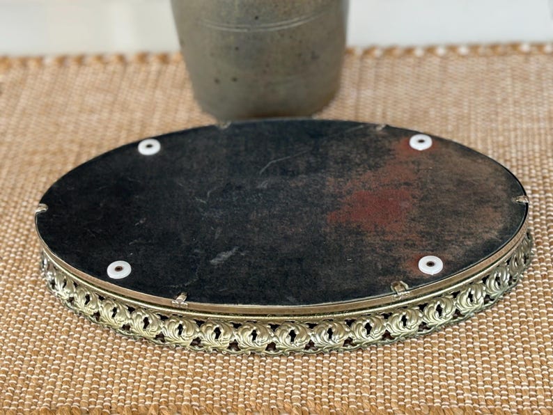 May include: An oval-shaped vanity tray featuring a dark, weathered backing with four white circular feet. The tray is enclosed by a decorative, perforated gold-toned metal rim with a repeating leaf-like pattern along the entire outer edge.