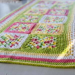 May include: A crocheted table runner with a green, white, yellow, and pink geometric pattern. The runner is made with a variety of stitches and has a fringed edge.