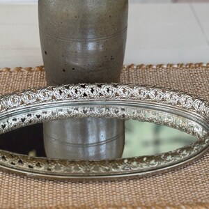May include: An oval silver-toned vanity tray featuring a reflective mirrored base and an intricate, raised metal gallery rim with a repeating openwork pattern. The tray has a polished metallic finish and a low-profile design for display.