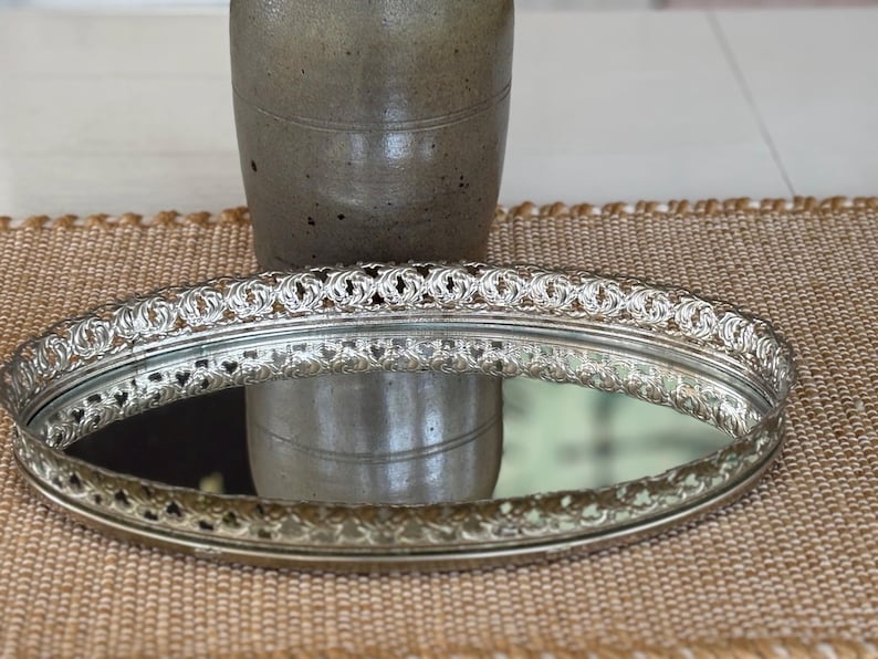 May include: An oval silver-toned vanity tray featuring a reflective mirrored base and an intricate, raised metal gallery rim with a repeating openwork pattern. The tray has a polished metallic finish and a low-profile design for display.