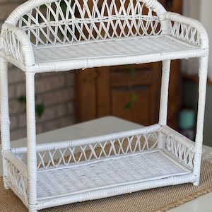 May include: A white wicker shelf with two tiers. The shelf has a curved back and is made of woven wicker. The shelf is designed to hold items and is suitable for display or storage. The shelf is painted white.