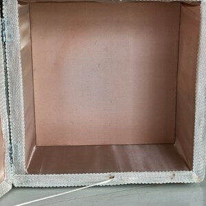 May include: A white fabric box with a pink satin lining. The box has a lace trim around the edges and a white string attached to the lid.