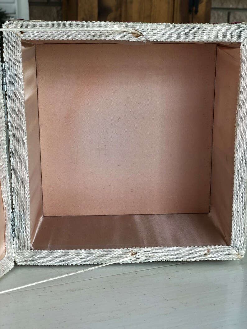 May include: A white fabric box with a pink satin lining. The box has a lace trim around the edges and a white string attached to the lid.