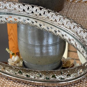 May include: An oval vanity tray with a reflective mirrored base and a raised, ornate silver-toned metal gallery rim featuring a repeating scrollwork pattern. The metallic edge has a decorative, pierced design that encircles the entire perimeter.
