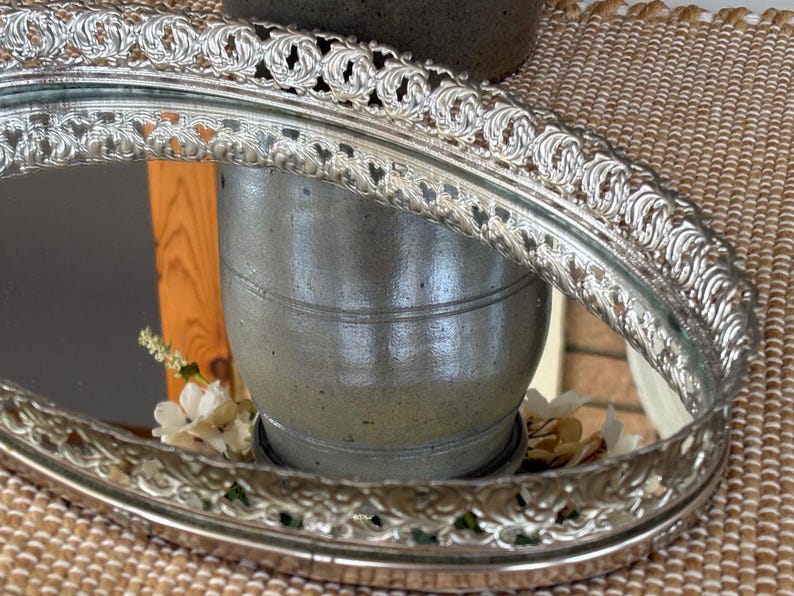 May include: An oval vanity tray with a reflective mirrored base and a raised, ornate silver-toned metal gallery rim featuring a repeating scrollwork pattern. The metallic edge has a decorative, pierced design that encircles the entire perimeter.