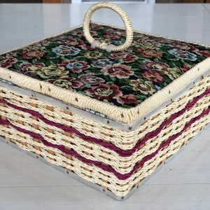 May include: A woven wicker box with a floral patterned fabric lid and a metal handle. The box has a burgundy trim and is closed.