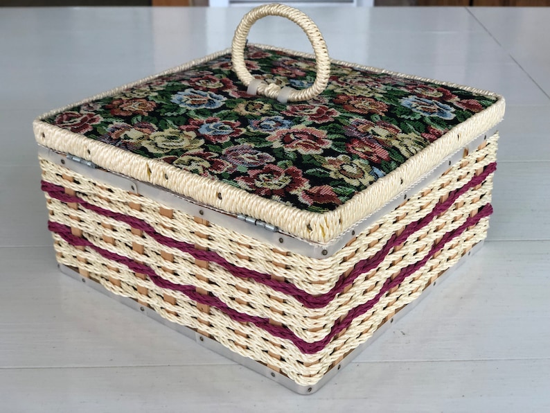May include: A woven wicker box with a floral patterned fabric lid and a metal handle. The box has a burgundy trim and is closed.
