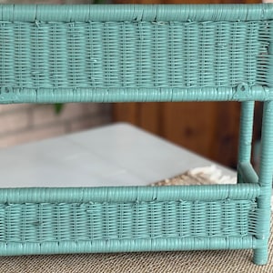 May include: A turquoise wicker shelf unit with a rectangular shape. The shelf has two levels and is supported by four legs. The wicker is woven in a tight pattern, and the unit appears to be painted. The background is blurred, but a neutral-colored surface is visible.