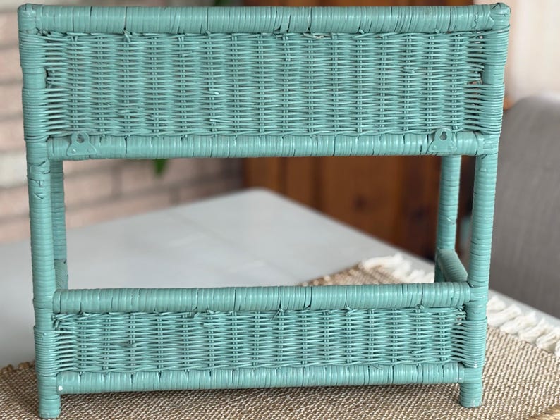 May include: A turquoise wicker shelf unit with a rectangular shape. The shelf has two levels and is supported by four legs. The wicker is woven in a tight pattern, and the unit appears to be painted. The background is blurred, but a neutral-colored surface is visible.