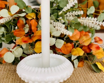 Può includere: Portacandele in vetro di latte bianco con una candela bianca. Il portacandele ha un design a perline e si trova su una superficie intrecciata marrone.