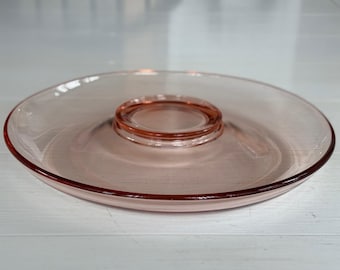 May include: A clear, pink glass serving dish with a circular, raised center section. The rim and center are a darker shade of pink. The dish is likely used for serving snacks or appetizers. The dish is on a white surface.