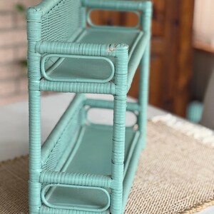 May include: A two-tiered, light teal wicker shelf. The shelf has a rectangular shape with rounded edges and a woven back. The shelves are supported by vertical posts and horizontal bars. The piece is painted in a solid, muted teal color.