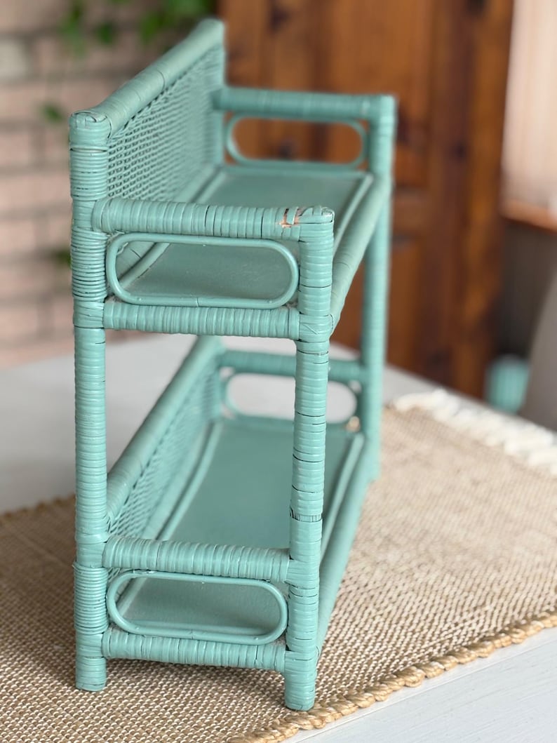 May include: A two-tiered, light teal wicker shelf. The shelf has a rectangular shape with rounded edges and a woven back. The shelves are supported by vertical posts and horizontal bars. The piece is painted in a solid, muted teal color.