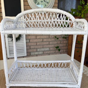 May include: A white wicker shelf with two levels. The shelf has a curved back and a woven design on the shelves and back. The shelf is made of white wicker and is suitable for displaying items.