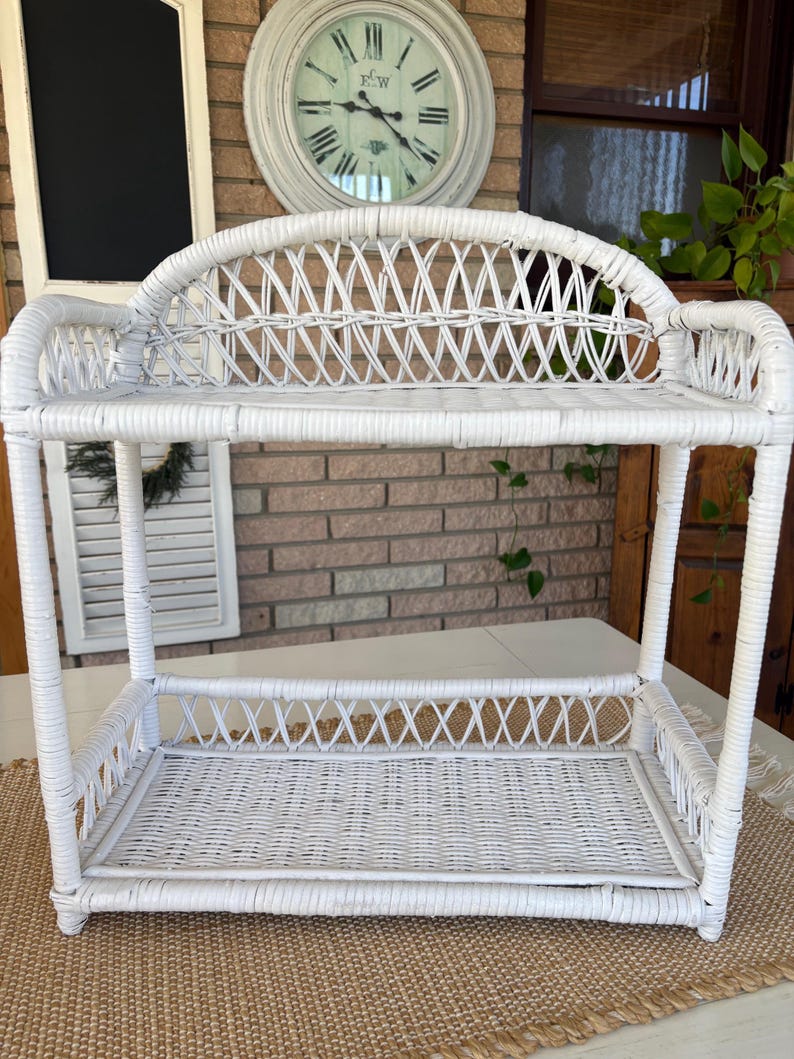 May include: A white wicker shelf with two levels. The shelf has a curved back and a woven design on the shelves and back. The shelf is made of white wicker and is suitable for displaying items.