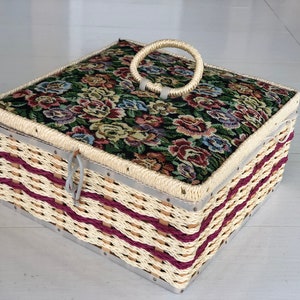 May include: A woven wicker storage box with a floral patterned fabric lid and a white plastic handle. The box has a burgundy and white striped design.