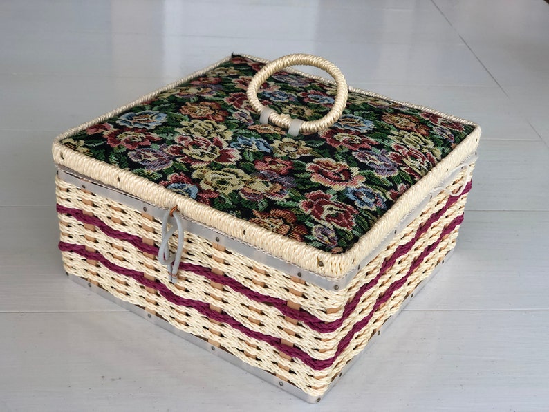 May include: A woven wicker storage box with a floral patterned fabric lid and a white plastic handle. The box has a burgundy and white striped design.