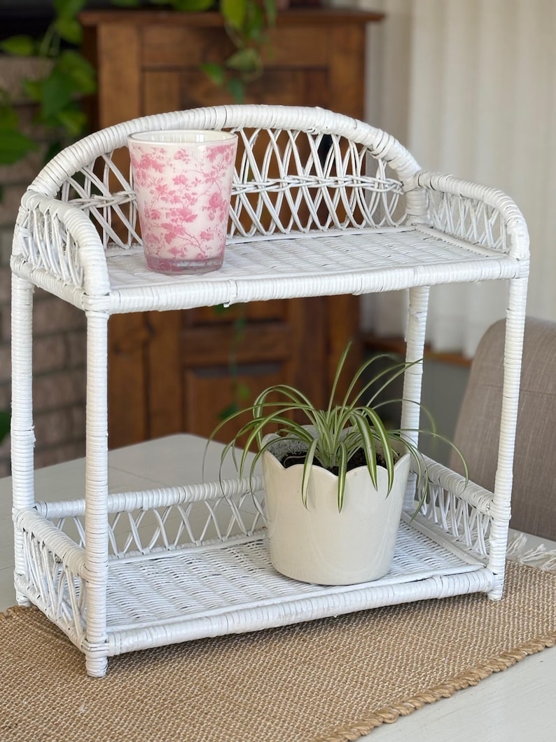 May include: A white wicker shelf unit with two tiers. A pink floral-patterned glass sits on the top shelf, and a white scalloped-edge pot with a green plant sits on the bottom shelf. The shelf unit is on a tan placemat.