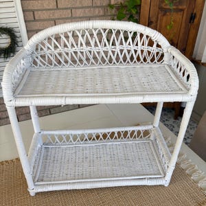 May include: A white wicker shelf with two levels. The top shelf has a curved back, and both shelves have a woven design. The shelf is made of white wicker and is suitable for displaying items. The shelf is on a neutral-colored rug.