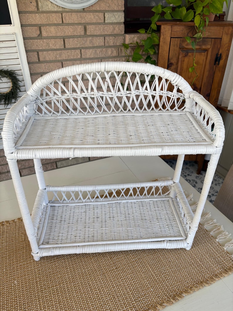 May include: A white wicker shelf with two levels. The top shelf has a curved back, and both shelves have a woven design. The shelf is made of white wicker and is suitable for displaying items. The shelf is on a neutral-colored rug.