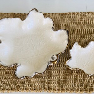 May include: Two white ceramic leaf-shaped serving dishes with gold trim. The larger dish is in the foreground and the smaller dish is behind it. Both dishes are on a brown woven surface.