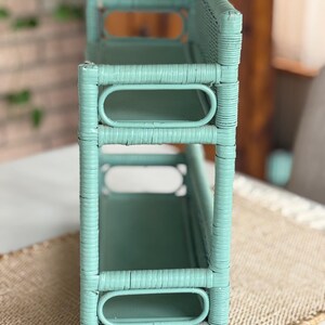 May include: A mint green, woven wicker shelf unit with two shelves and a back. The shelf has an open design with oval cutouts and a rectangular shape. The unit is painted in a solid, light green color.