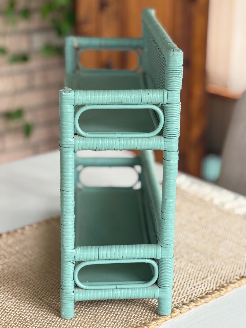 May include: A mint green, woven wicker shelf unit with two shelves and a back. The shelf has an open design with oval cutouts and a rectangular shape. The unit is painted in a solid, light green color.