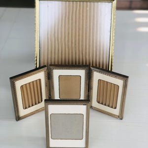 May include: Set of five gold-toned picture frames with ornate detailing. The frames are arranged in a cluster, with a larger frame at the top and four smaller frames below. The frames are empty and ready to display photos.