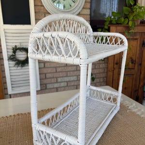 May include: A white, two-tiered wicker shelf with a curved top and rectangular shelves. The shelf has a woven design and is suitable for displaying items. The shelf is made of white wicker and is suitable for a bathroom or kitchen.