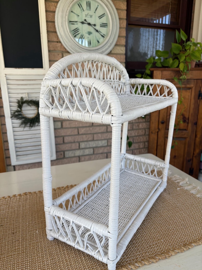 May include: A white, two-tiered wicker shelf with a curved top and rectangular shelves. The shelf has a woven design and is suitable for displaying items. The shelf is made of white wicker and is suitable for a bathroom or kitchen.