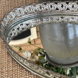 May include: An oval silver-toned vanity tray featuring a raised, ornate filigree border with intricate scrollwork patterns. The center of the tray consists of a reflective mirror surface that displays a reflection of a gray ceramic vessel.