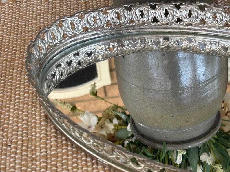 May include: An oval silver-toned vanity tray featuring a raised, ornate filigree border with intricate scrollwork patterns. The center of the tray consists of a reflective mirror surface that displays a reflection of a gray ceramic vessel.