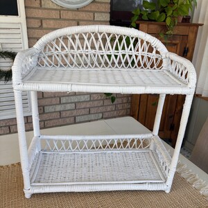 May include: A white wicker shelf with two levels. The top shelf has a curved back. The shelves and supports are made of woven wicker. The shelf is on a white table with a woven mat underneath.