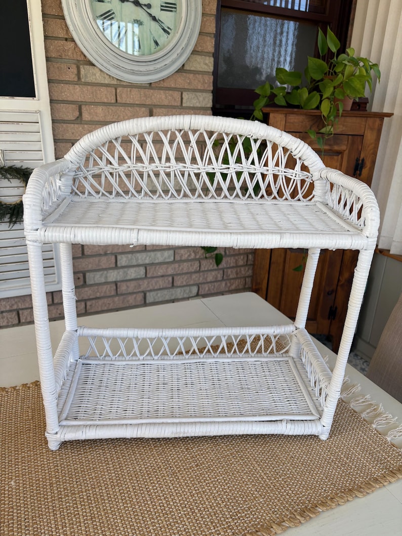 May include: A white wicker shelf with two levels. The top shelf has a curved back. The shelves and supports are made of woven wicker. The shelf is on a white table with a woven mat underneath.