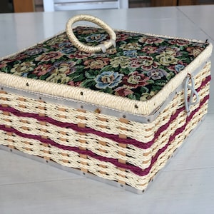 May include: A woven wicker sewing box with a floral patterned fabric lid and a white plastic handle. The box has a burgundy trim and metal accents.
