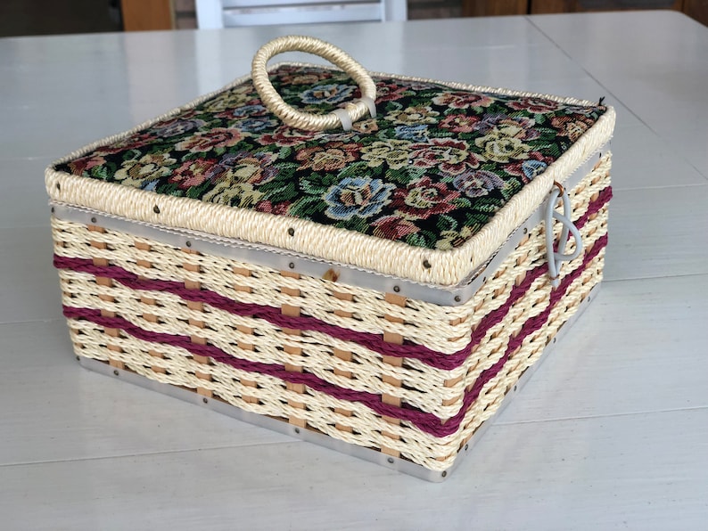 May include: A woven wicker sewing box with a floral patterned fabric lid and a white plastic handle. The box has a burgundy trim and metal accents.