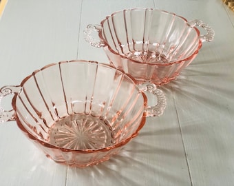 May include: Two pink glass bowls with fluted sides and decorative handles. The bowls are empty and sitting on a white surface.