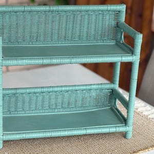 May include: A teal-colored, two-tiered wicker shelf. The shelf has a rectangular shape with a woven design on the back and between the shelves. The shelves are flat and the frame is made of rounded wicker. The shelf is designed for storage or display.