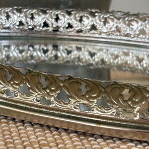 May include: An oval vanity tray with a mirrored base and a raised, metallic silver-toned gallery rail. The perimeter features an intricate, repeating openwork scroll pattern that encircles the reflective surface, creating a decorative border.