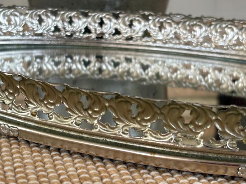 May include: An oval vanity tray with a mirrored base and a raised, metallic silver-toned gallery rail. The perimeter features an intricate, repeating openwork scroll pattern that encircles the reflective surface, creating a decorative border.