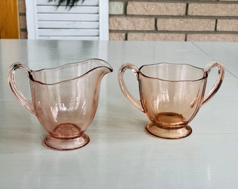 May include: A vintage pink glass creamer and sugar bowl set. The creamer has a single handle and the sugar bowl has two handles.