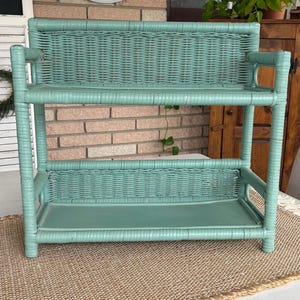 May include: A vintage, two-tiered wicker shelf painted in a soft teal color. The shelf has a rectangular shape with woven detailing on the shelves and back. The shelf is supported by rounded posts and is placed on a woven mat.