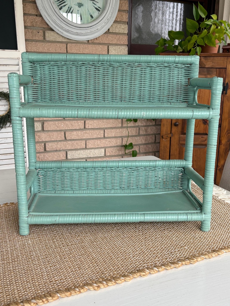 May include: A vintage, two-tiered wicker shelf painted in a soft teal color. The shelf has a rectangular shape with woven detailing on the shelves and back. The shelf is supported by rounded posts and is placed on a woven mat.