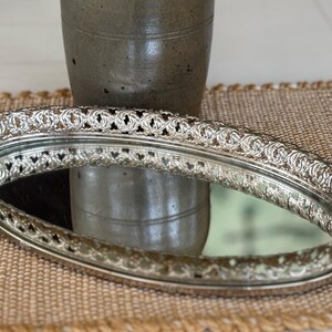 May include: An oval vanity tray featuring a reflective mirrored base and a raised, intricate silver-toned metal gallery rim with a repeating openwork scroll pattern. The tray has a polished metallic finish and a classic, elongated oval shape.