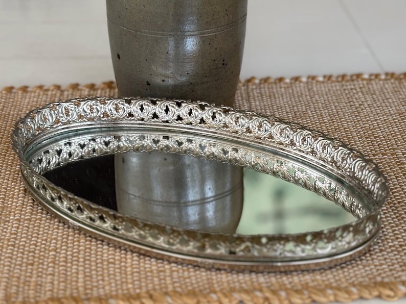 May include: An oval vanity tray featuring a reflective mirrored base and a raised, intricate silver-toned metal gallery rim with a repeating openwork scroll pattern. The tray has a polished metallic finish and a classic, elongated oval shape.
