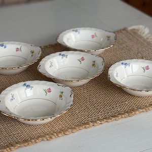 May include: Five white porcelain bowls with scalloped edges and gold rims. Each bowl is decorated with small floral patterns in blue, pink, and yellow. The bowls are arranged on a woven beige placemat.