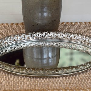 May include: An oval-shaped silver-toned vanity tray featuring a reflective mirrored base and an intricate, raised metal border with a repeating openwork pattern along the entire perimeter of the tray's edge.