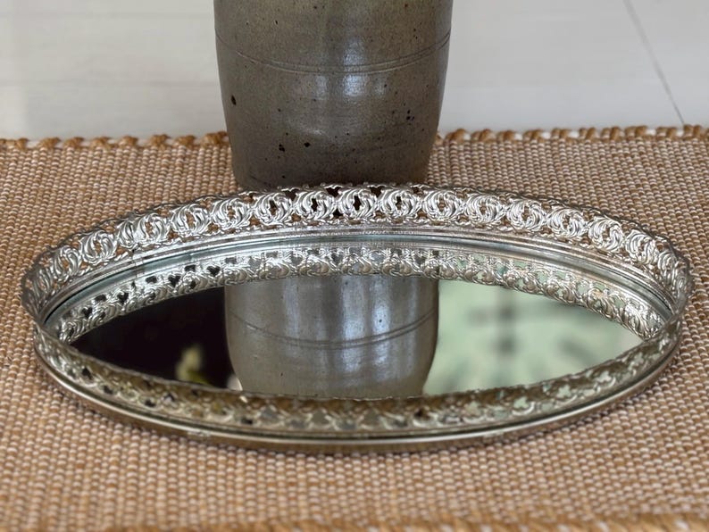 May include: An oval-shaped silver-toned vanity tray featuring a reflective mirrored base and an intricate, raised metal border with a repeating openwork pattern along the entire perimeter of the tray's edge.