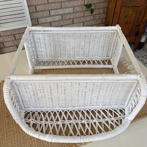 May include: A white wicker wall shelf with a curved bottom and two basket-like compartments. The shelf is made of woven wicker and has a clean, bright appearance. The shelf is designed to be mounted on a wall and is suitable for storage or display.
