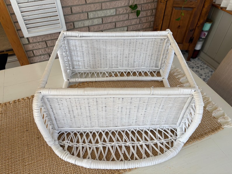 May include: A white wicker wall shelf with a curved bottom and two basket-like compartments. The shelf is made of woven wicker and has a clean, bright appearance. The shelf is designed to be mounted on a wall and is suitable for storage or display.